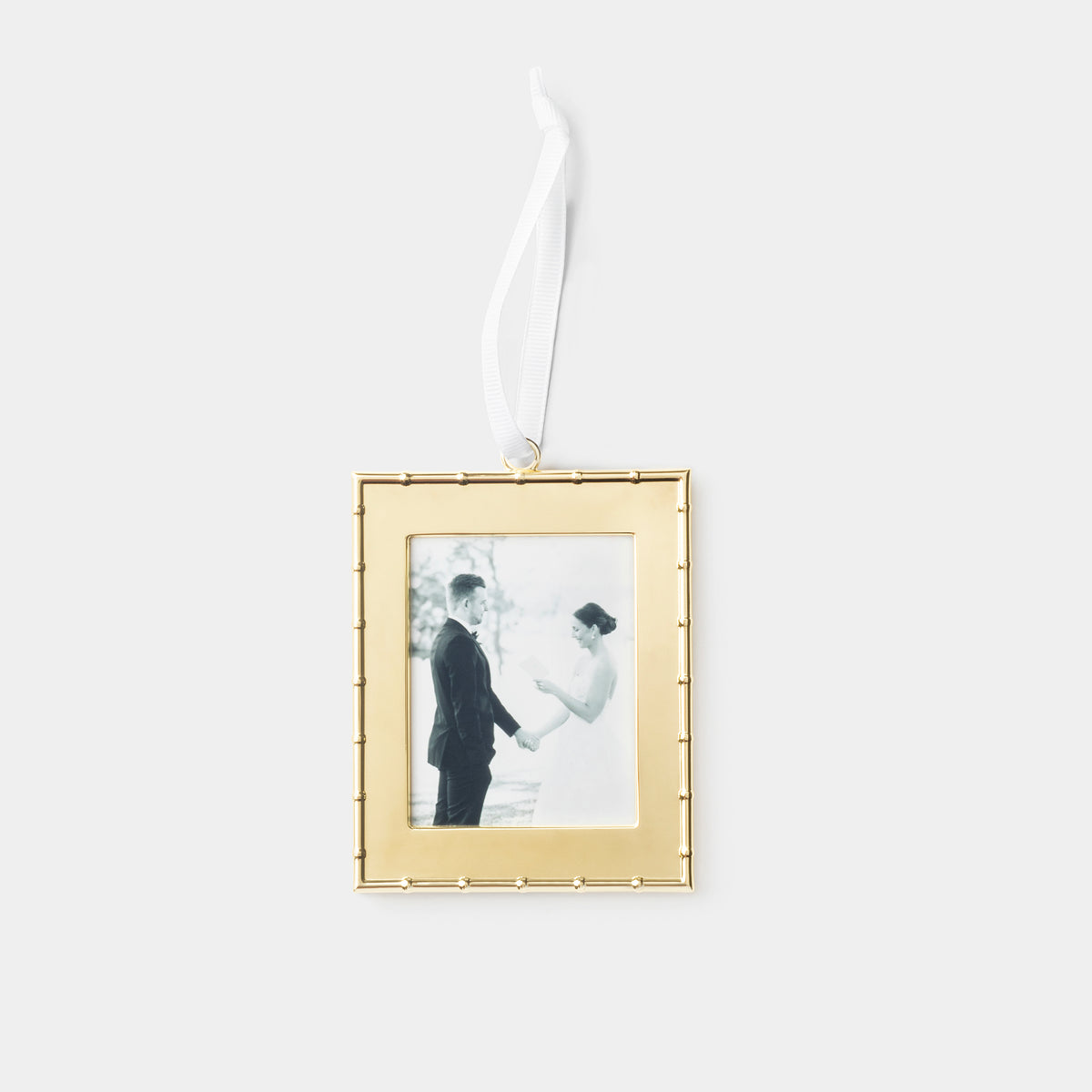 A rectangular gold picture frame ornament with a white hanging ribbon, featuring a black-and-white wedding photo of a couple, shown against a light gray background.